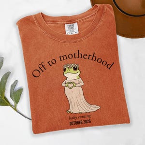 May include: Burnt orange t-shirt with the text "Off to motherhood" and a cartoon frog wearing a floral crown and dress, forming a heart with its hands. The text "baby coming OCTOBER 2026" is below the frog.