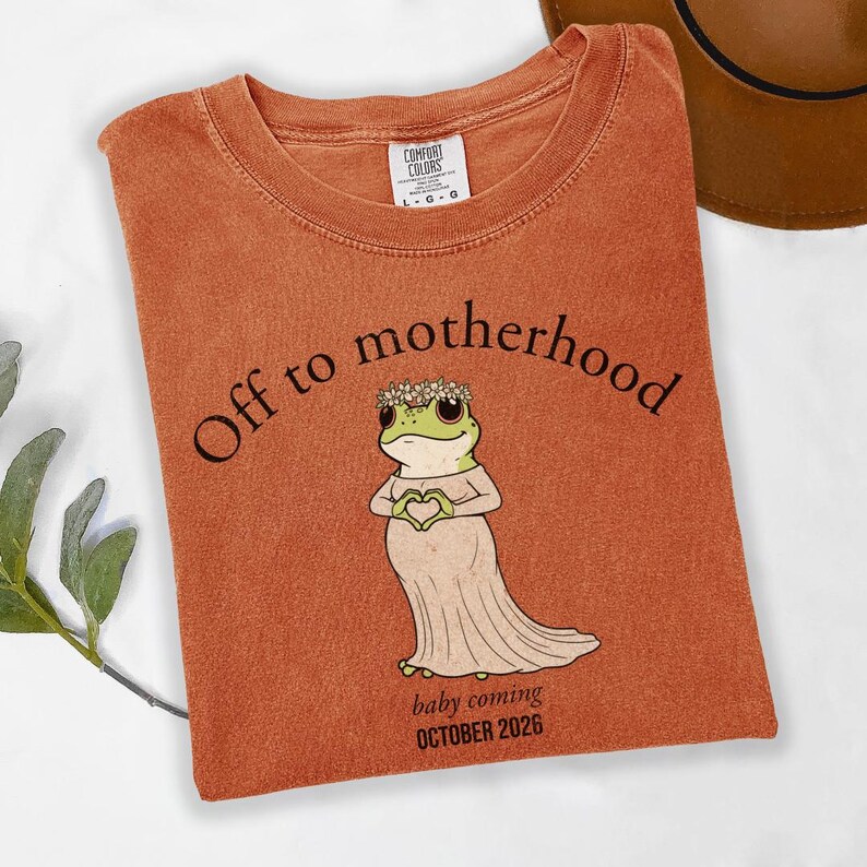 May include: Burnt orange t-shirt with the text "Off to motherhood" and a cartoon frog wearing a floral crown and dress, forming a heart with its hands. The text "baby coming OCTOBER 2026" is below the frog.