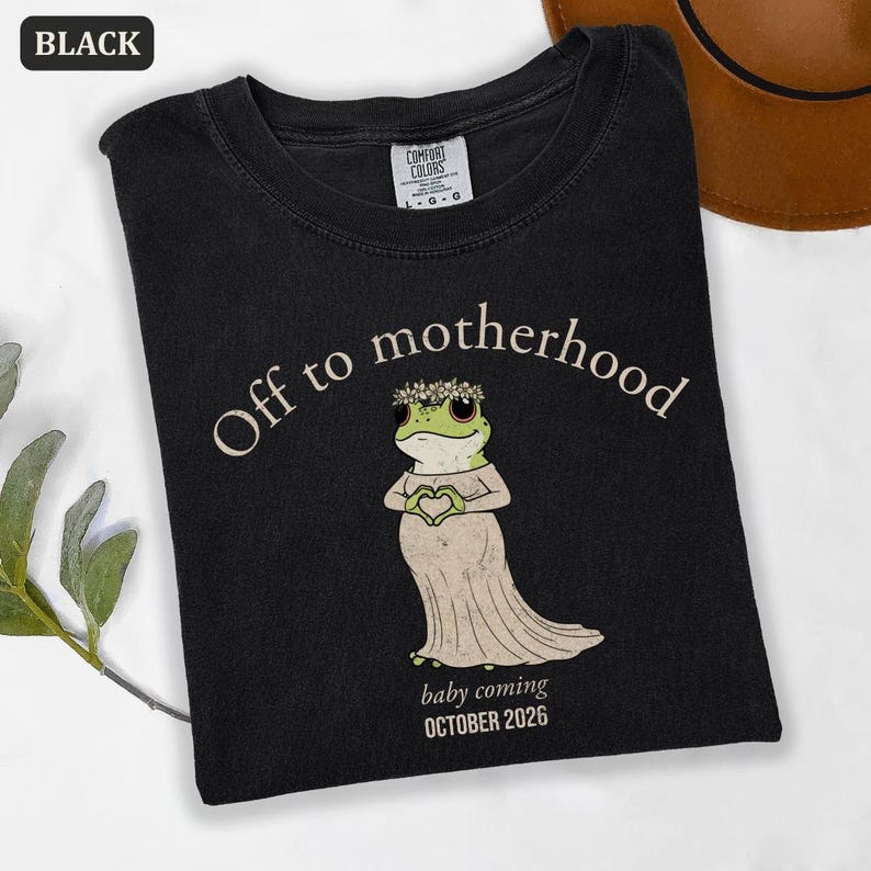May include: Black t-shirt with the text "Off to motherhood" and a cartoon frog wearing a floral crown and dress, holding a heart. The text "baby coming OCTOBER 2026" is below the frog. The shirt is labeled "BLACK" in the upper left corner.