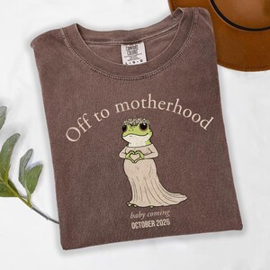 May include: Brown t-shirt with the text "Off to motherhood" and a cartoon frog wearing a floral crown and dress. The text "baby coming OCTOBER 2026" is also printed on the shirt. The shirt is made by Comfort Colors.