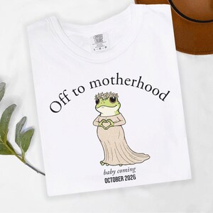 May include: White t-shirt with the text "Off to motherhood" above a cartoon frog wearing a dress and flower crown, holding a heart. The text "baby coming OCTOBER 2026" is below the frog.