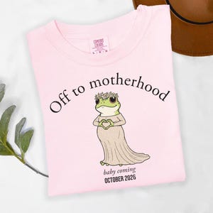 May include: A light pink t-shirt with the words "Off to motherhood" and a cartoon frog wearing a dress and flower crown. The text "baby coming OCTOBER 2026" is below the frog. The shirt is a Comfort Colors brand.