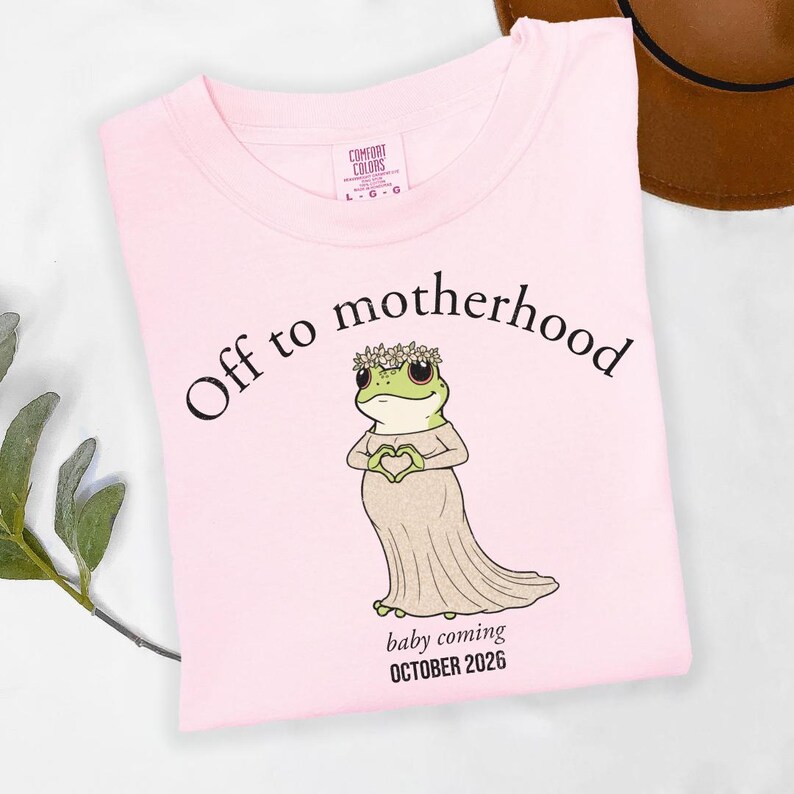 May include: A light pink t-shirt with the words "Off to motherhood" and a cartoon frog wearing a dress and flower crown. The text "baby coming OCTOBER 2026" is below the frog. The shirt is a Comfort Colors brand.