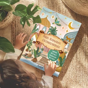Può includere: Un libro di attività per bambini intitolato "Mein Ramadan Entdeckerbuch" con illustrazioni colorate di lanterne, stelle e una luna crescente. Il libro è aperto, con le mani di un bambino visibili, su una superficie tessuta.