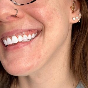 May include: Close-up of a person's smiling mouth with bright white teeth. A small gem is affixed to one tooth. The person is wearing a nose ring and glasses. The ear has multiple earrings, including a gold spiral and several small, sparkly studs.