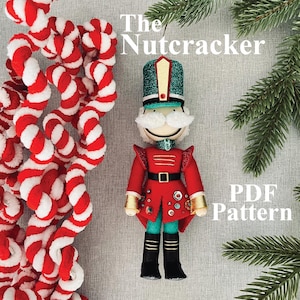 May include: A fabric nutcracker ornament with a green glitter hat, red coat, and black boots. The figure has a white mustache and is decorated with small buttons. Text reads The Nutcracker PDF Pattern. A red and white twisted rope is on the left.