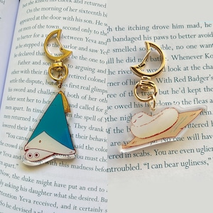 May include: Two acrylic keychains with gold-colored hardware. One features a blue and white hat with a tassel, the other depicts a sleeping creature on a rug. The keychains are set against the backdrop of an open book.
