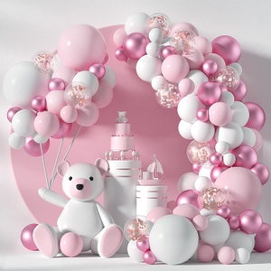 May include: A whimsical display featuring a large white teddy bear, a tiered cake, and a sailboat figurine. The scene is adorned with a balloon arch in shades of pink, white, and clear balloons with confetti, creating a festive atmosphere.