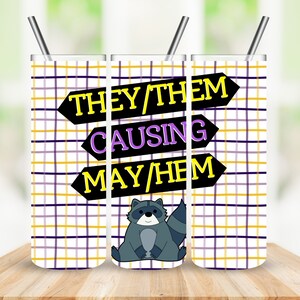 May include: A stainless steel tumbler with a plaid pattern in yellow, purple, and white. The tumbler features the text "THEY/THEM CAUSING MAYHEM" in black arrows, and a cartoon raccoon. Includes a silver straw.