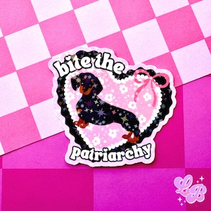 May include: A sticker featuring a dachshund dog inside a pink heart with a floral pattern. The sticker has the text "bite the Patriarchy" and a pink bow. The background is a pink and white checkered pattern.