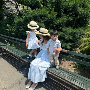 K&ouml;nnte beinhalten: Eine dreik&ouml;pfige Familie in passenden blau-wei&szlig; karierten Outfits. Die Erwachsenen tragen Strohh&uuml;te, das Kind ein blau-wei&szlig; gestreiftes Hemd. Sie sitzen auf einer Holzbank in einem Park.