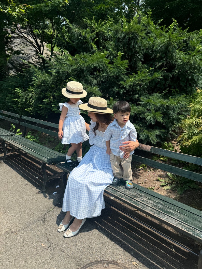 K&ouml;nnte beinhalten: Eine dreik&ouml;pfige Familie in passenden blau-wei&szlig; karierten Outfits. Die Erwachsenen tragen Strohh&uuml;te, das Kind ein blau-wei&szlig; gestreiftes Hemd. Sie sitzen auf einer Holzbank in einem Park.