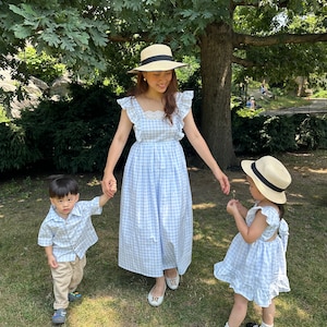 K&ouml;nnte beinhalten: Eine dreik&ouml;pfige Familie tr&auml;gt passende blaue und wei&szlig;e Gingham-Kleider und Hemden. Die Frau tr&auml;gt einen Strohhut und h&auml;lt die Hand eines kleinen Jungen. Das M&auml;dchen h&auml;lt eine kleine Blume und tr&auml;gt einen Strohhut. Sie stehen auf einem Rasen.
