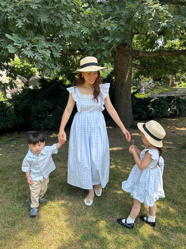 K&ouml;nnte beinhalten: Eine dreik&ouml;pfige Familie tr&auml;gt passende blaue und wei&szlig;e Gingham-Kleider und Hemden. Die Frau tr&auml;gt einen Strohhut und h&auml;lt die Hand eines kleinen Jungen. Das M&auml;dchen h&auml;lt eine kleine Blume und tr&auml;gt einen Strohhut. Sie stehen auf einem Rasen.