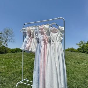 May include: Five white cotton dresses with different patterns hanging on a white rack against a blue sky. The dresses have ruffled straps and are all different lengths. The dresses are in a variety of patterns, including gingham, polka dots, and plain white.