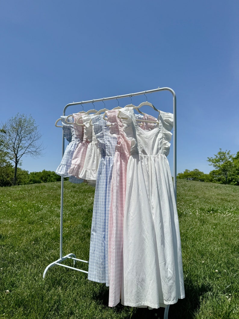 May include: Five white cotton dresses with different patterns hanging on a white rack against a blue sky. The dresses have ruffled straps and are all different lengths. The dresses are in a variety of patterns, including gingham, polka dots, and plain white.