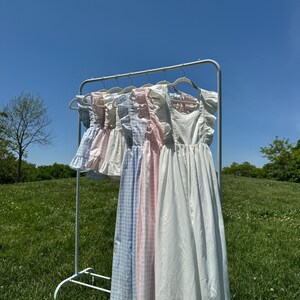 K&ouml;nnte beinhalten: Ein Gestell mit f&uuml;nf wei&szlig;en, rosa und blauen Gingham-Kleidern mit R&uuml;schenbesatz, die an einem Metallgestell vor einem Hintergrund aus blauem Himmel und gr&uuml;nem Gras h&auml;ngen.