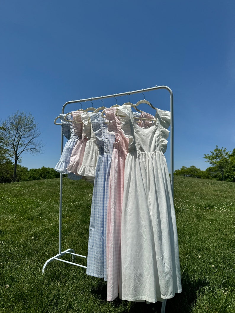 K&ouml;nnte beinhalten: Ein Gestell mit f&uuml;nf wei&szlig;en, rosa und blauen Gingham-Kleidern mit R&uuml;schenbesatz, die an einem Metallgestell vor einem Hintergrund aus blauem Himmel und gr&uuml;nem Gras h&auml;ngen.