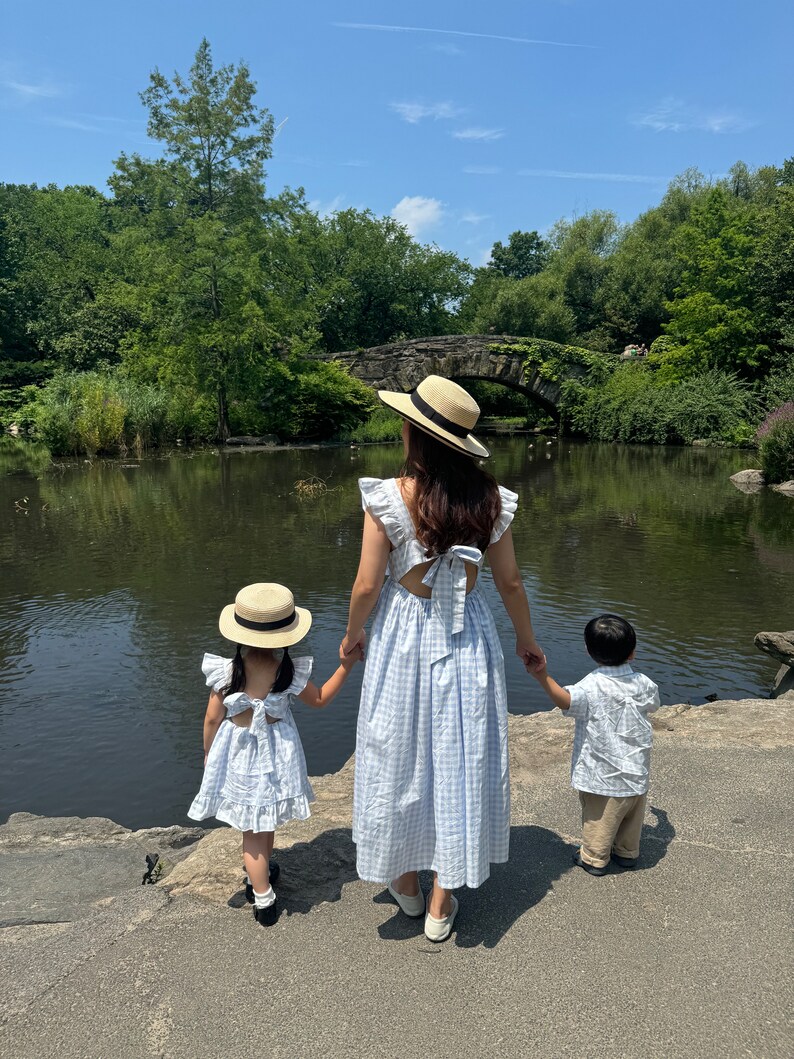 K&ouml;nnte beinhalten: Eine dreik&ouml;pfige Familie in passenden blau-wei&szlig;en Gingham-Kleidern und einem wei&szlig;en Hemd steht am Rande eines Teichs in einem Park. Die Frau tr&auml;gt einen Strohhut mit schwarzem Band, die Kinder tragen Strohh&uuml;te mit wei&szlig;en B&auml;ndern. Sie schauen alle auf den Teich.