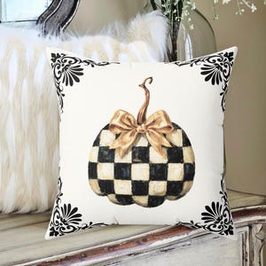 May include: Square decorative pillow with a cream background and a watercolor pumpkin design. The pumpkin is black and cream checkered with a gold bow. Black floral corner accents frame the design. The pillow is on a white dresser.