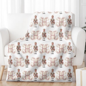May include: White throw blanket with a repeating pattern of a nutcracker soldier and the letter H in a pink and gold design.