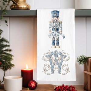May include: White kitchen towel featuring a blue and white nutcracker design and a monogram. The nutcracker is in a classic uniform with a hat and trumpet. The monogram is a stylized "W" in blue and tan.