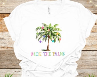 May include: White t-shirt with a green palm tree decorated with colorful ornaments. The text "Deck the Palms" is printed in rainbow colors below the tree.