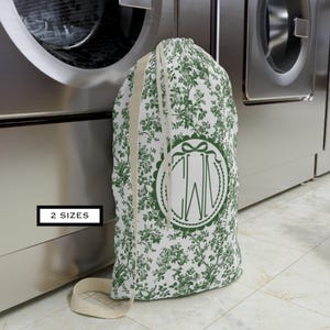 May include: A white and green floral print laundry bag with a drawstring closure and a beige strap. The bag features a green monogram in a decorative oval. The text "2 SIZES" is in a black box.