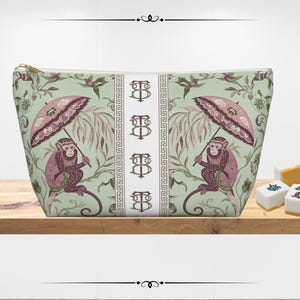 Puede incluir: Bolsa de cosméticos rectangular con cremallera dorada. La bolsa presenta un patrón repetido de monos con paraguas, sobre un fondo verde claro con motivos florales y de hojas. Una franja blanca con la letra "B" está en el centro.