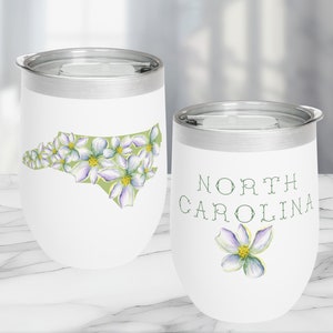 May include: Two white stainless steel wine tumblers with a North Carolina theme. One tumbler features an outline of the state of North Carolina with white flowers. The other tumbler has the words "North Carolina" and a single white flower.