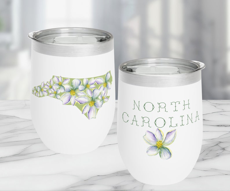 May include: Two white stainless steel wine tumblers with a North Carolina theme. One tumbler features an outline of the state of North Carolina with white flowers. The other tumbler has the words "North Carolina" and a single white flower.