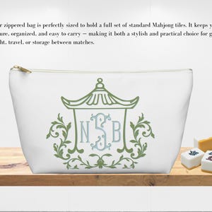 May include: White zippered bag with a green and blue decorative design. The bag features a gold zipper and is designed to hold Mahjong tiles. The bag is on a wooden surface with Mahjong tiles.