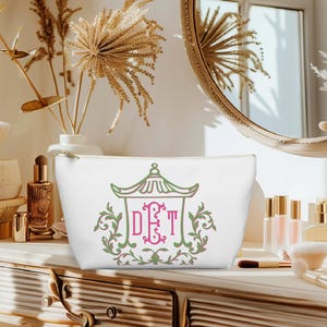 May include: White cosmetic bag with a gold zipper, featuring a green and pink pagoda design with the initials DET. The bag is sitting on a wooden vanity with makeup brushes and perfume bottles.