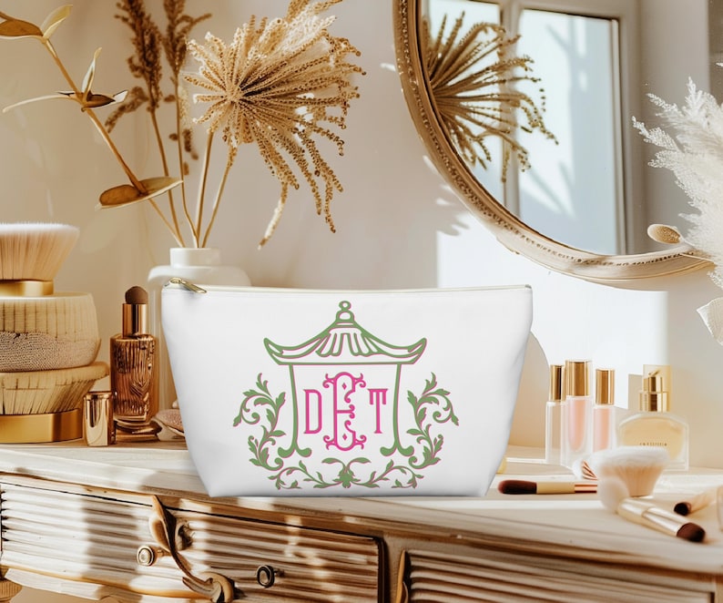 May include: White cosmetic bag with a gold zipper, featuring a green and pink pagoda design with the initials DET. The bag is sitting on a wooden vanity with makeup brushes and perfume bottles.