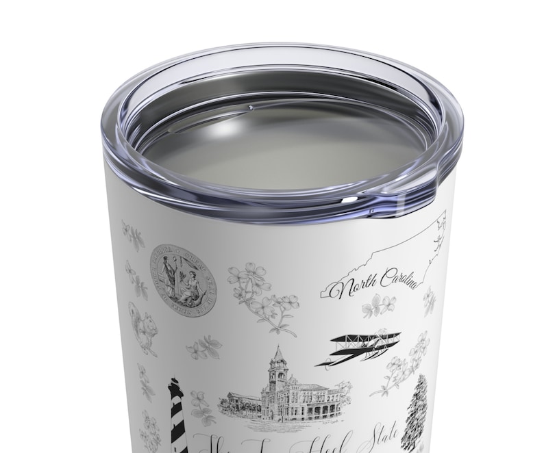 May include: White stainless steel tumbler with a black and white design featuring various North Carolina landmarks, including the state seal, a lighthouse, a building, and an aeroplane. The text "North Carolina" is visible on the tumbler.