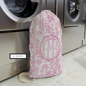 May include: A white and pink floral laundry bag with a drawstring closure and a beige strap. The bag features a monogram in a decorative circle. The text "2 SIZES" is in a black rectangle.