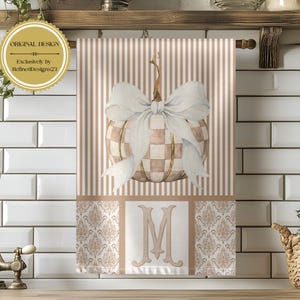 May include: A rectangular kitchen towel featuring a beige and white striped background with a central watercolor-style pumpkin decorated with a large white bow. The bottom section displays a beige damask pattern and a large, centered letter M.