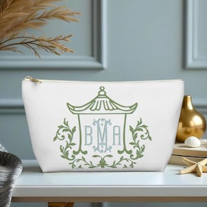 May include: White cosmetic bag with a gold zipper, featuring a green and blue pagoda design with the initials BMA. The design is surrounded by green floral elements. The bag is on a white surface.