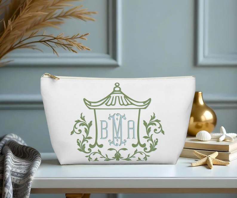 May include: White cosmetic bag with a gold zipper, featuring a green and blue pagoda design with the initials BMA. The design is surrounded by green floral elements. The bag is on a white surface.
