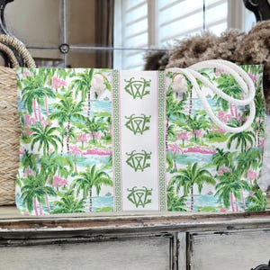 May include: A large tote bag with a tropical print featuring palm trees, pink accents, and a blue water scene. The bag has white rope handles and a white center panel with green monogrammed emblems.