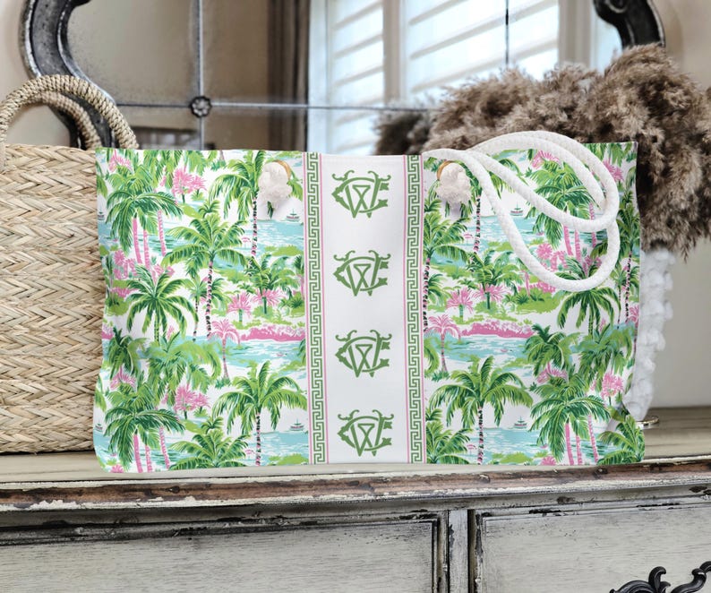 May include: A large tote bag with a tropical print featuring palm trees, pink accents, and a blue water scene. The bag has white rope handles and a white center panel with green monogrammed emblems.