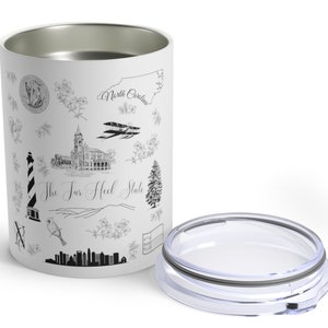 May include: White stainless steel tumbler with a black and white design featuring various landmarks and symbols of North Carolina, including the state outline, a lighthouse, a building, a bird, and the text "The Tar Heel State". The tumbler has a clear lid.