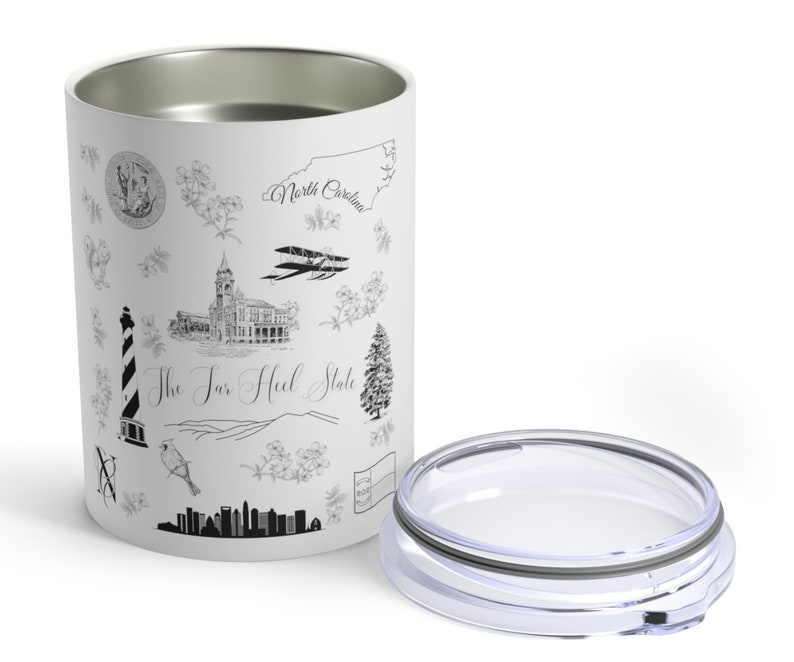 May include: White stainless steel tumbler with a black and white design featuring various landmarks and symbols of North Carolina, including the state outline, a lighthouse, a building, a bird, and the text "The Tar Heel State". The tumbler has a clear lid.