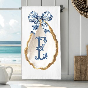 May include: White kitchen towel with a blue floral bow and an oyster shell design. The shell has a gold border and a blue letter "E" in the center. The towel is hanging on a silver bar.