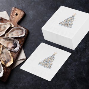 May include: Two white paper napkins with a Christmas tree design made of oysters. The tree is topped with a gold star. The napkins are on a dark gray surface.