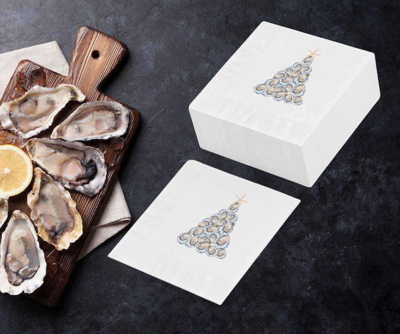 May include: Two white paper napkins with a Christmas tree design made of oysters. The tree is topped with a gold star. The napkins are on a dark gray surface.