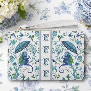 May include: A white clutch with a whimsical design featuring monkeys holding umbrellas, surrounded by blue and green floral and foliage patterns. The clutch has a white wrist strap and a gold zipper.