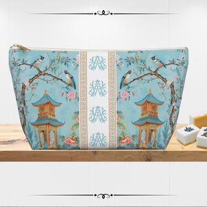 May include: A light blue cosmetic bag with a gold zipper, featuring a pagoda and bird design. The bag has a floral and branch pattern with a central white panel. Mahjong tiles are on a wooden shelf.
