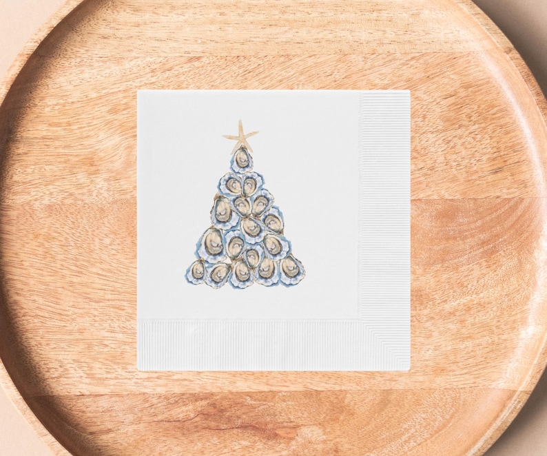 May include: A white paper napkin with a blue and white oyster shell Christmas tree design. The tree is topped with a gold starfish.