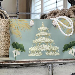 K&ouml;nnte beinhalten: Hellblaue Tragetasche mit wei&szlig;em Seilgriff. Die Tasche hat ein Blumendesign mit wei&szlig;en Lotusbl&uuml;ten und gr&uuml;nen Bl&auml;ttern. Goldene Palmwedel sind ebenfalls auf der Tasche aufgedruckt.